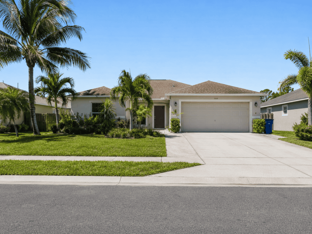 Single-story suburban Florida home with a two-car garage, palm trees, tropical landscaping, green lawn, and wide driveway under a clear blue sky.