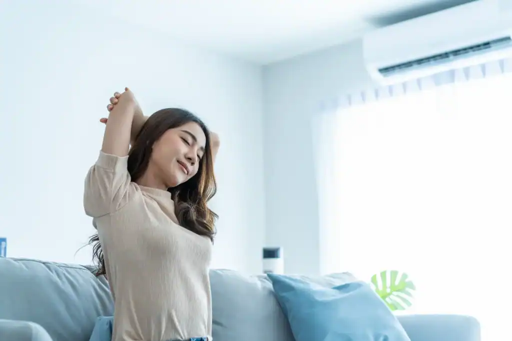 Woman stretching on a couch in a bright living room. The setting includes a white wall, an air conditioning unit.
