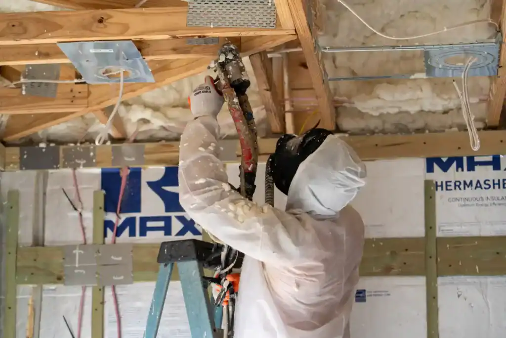 A professional in full body gear standing on a ladder spray foaming a new construction build of a house.