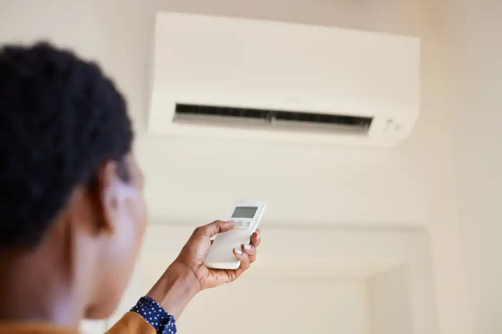 From a low angle, a person indoors holds a white remote control up towards a wall-mounted air conditioning unit. The person has short dark hair and is wearing a tan-colored jacket with a dark blue collared shirt with white polka dots underneath. The unit and the wall are both white. A doorway can be seen in the background.