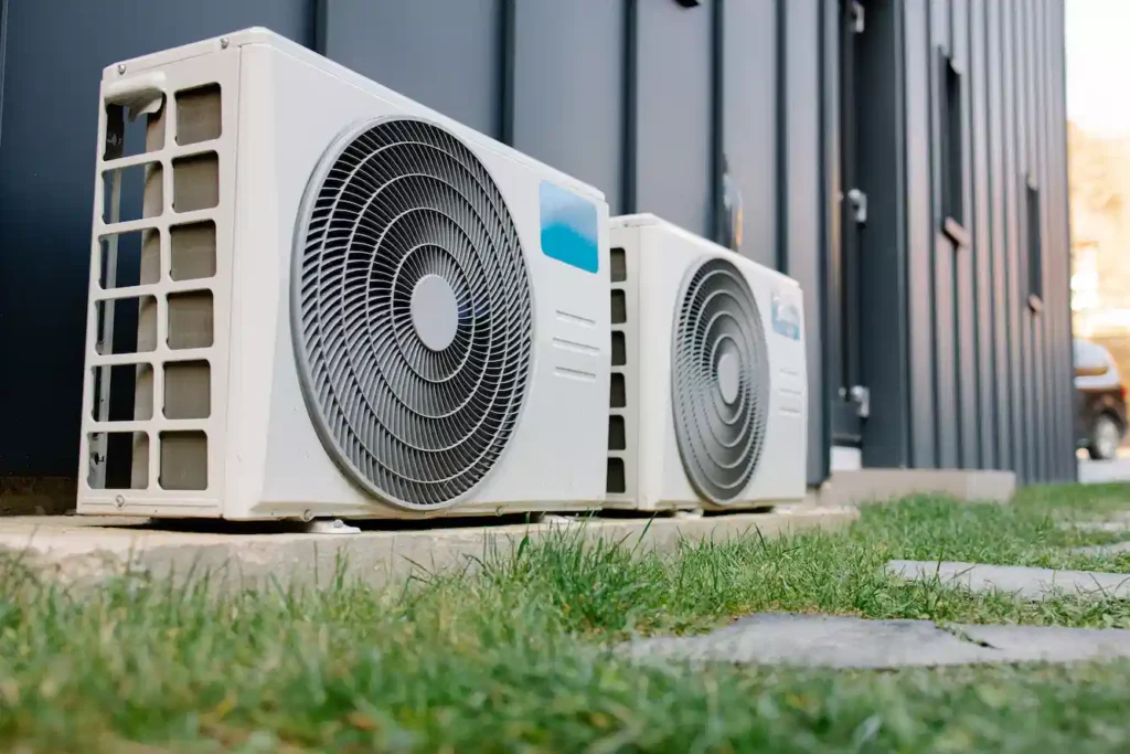 A view of two air conditioning units, situated on a concrete pad outside of a grey-paneled building